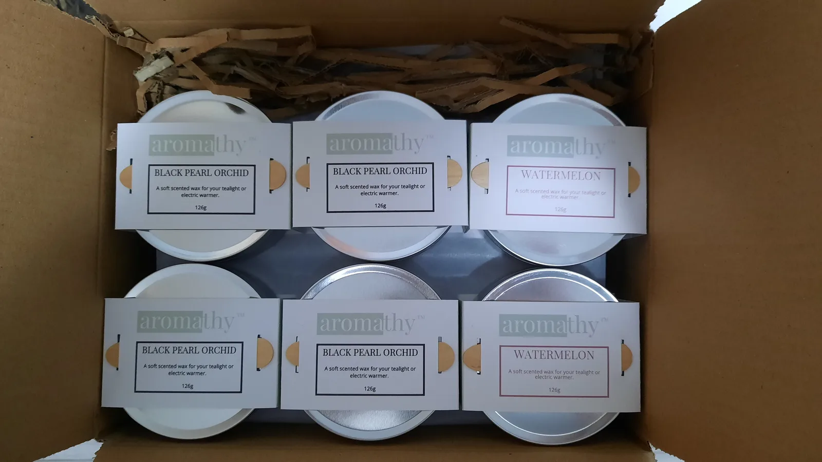 Six tins of Aromathy wax melts packed in a box with shredded paper, labelled Black Pearl Orchid and Watermelon